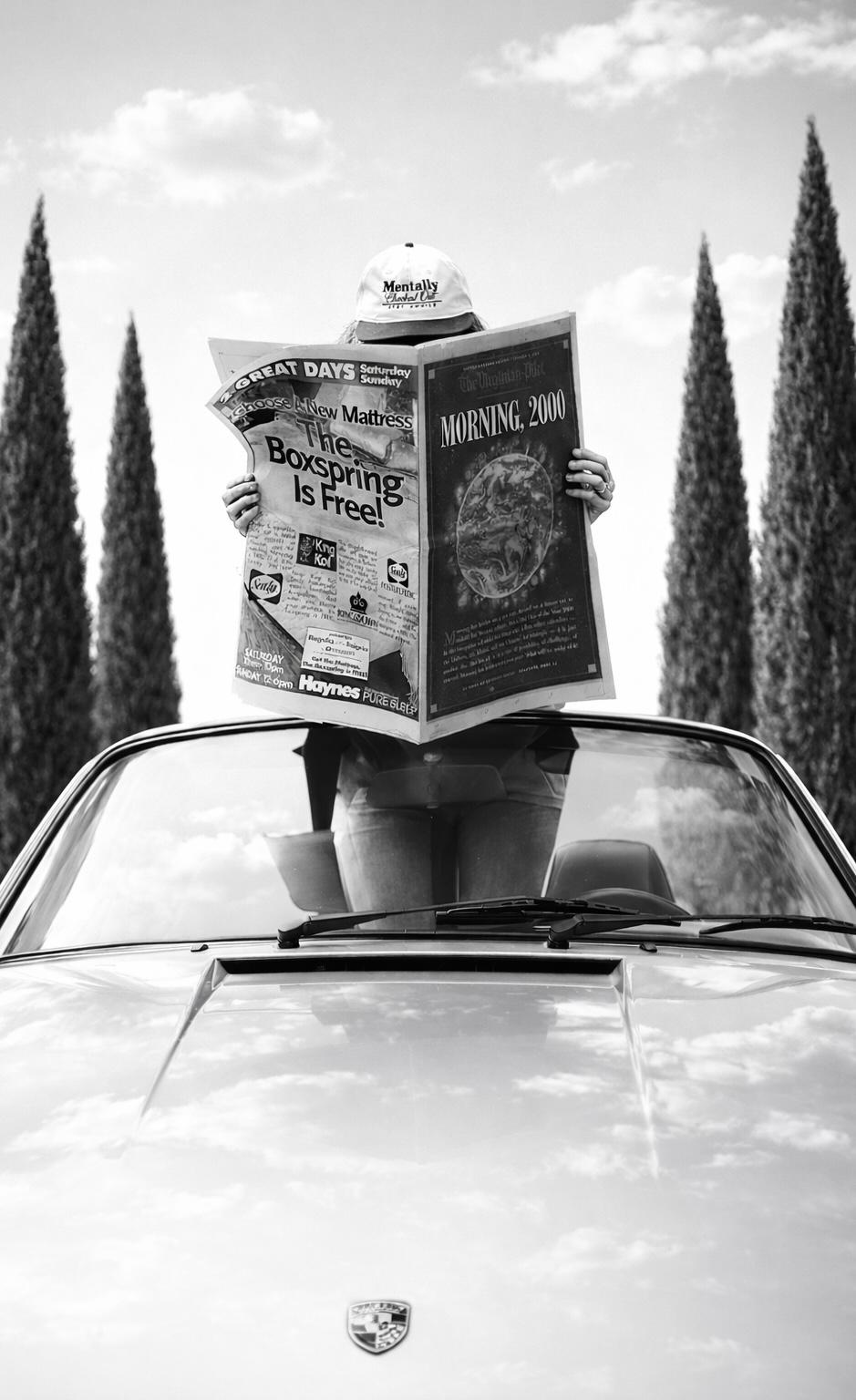 A person standing through a car's sunroof holding up a newspaper, framed by tall cypress trees.