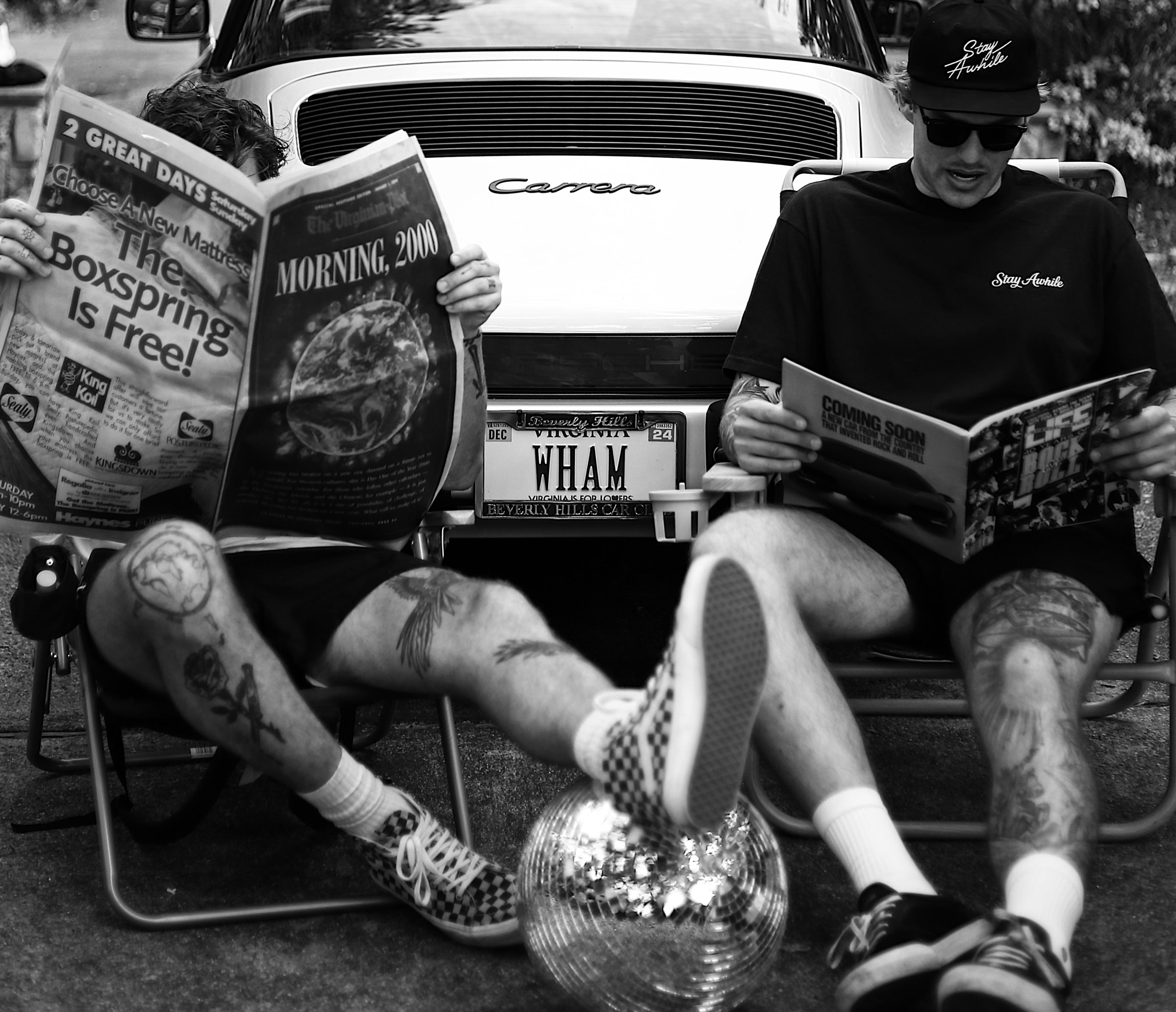 Two people reading newspapers in lawn chairs behind a white Porsche Carrera, a mirrorball resting on the asphalt between them.
